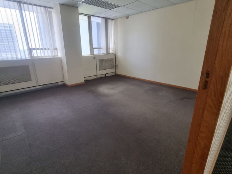 An empty commercial office space featuring a suspended ceiling, recessed lighting, carpeted flooring, and built-in HVAC units with large windows.