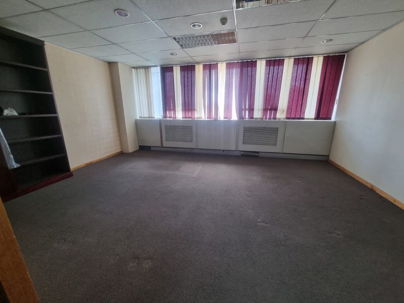 An empty commercial office space featuring a suspended ceiling, built-in HVAC units, and large windows with vertical blinds.
