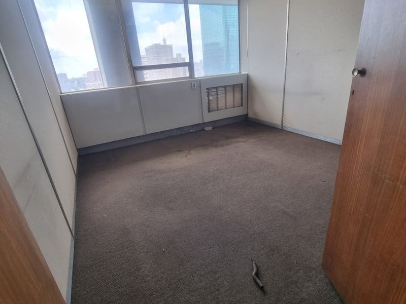 An empty, dated commercial office space featuring carpet flooring, partition walls, and large windows with city views, requiring cosmetic refurbishment.