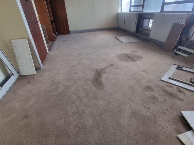 Vacant commercial office space featuring large windows, suspended ceiling, and built-in cabinetry, currently requiring significant refurbishment and flooring replacement.