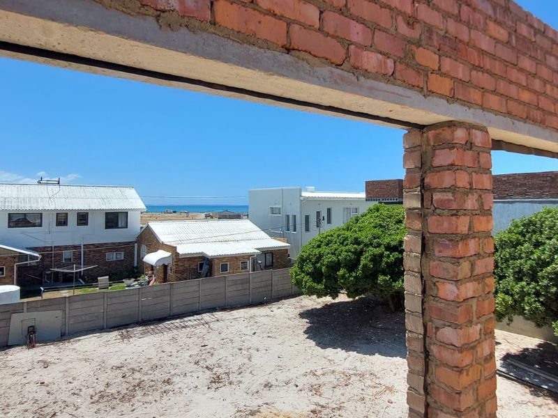 An unfinished construction site showing exposed brickwork and a concrete lintel, overlooking a rear yard with a view of neighboring properties and the ocean.