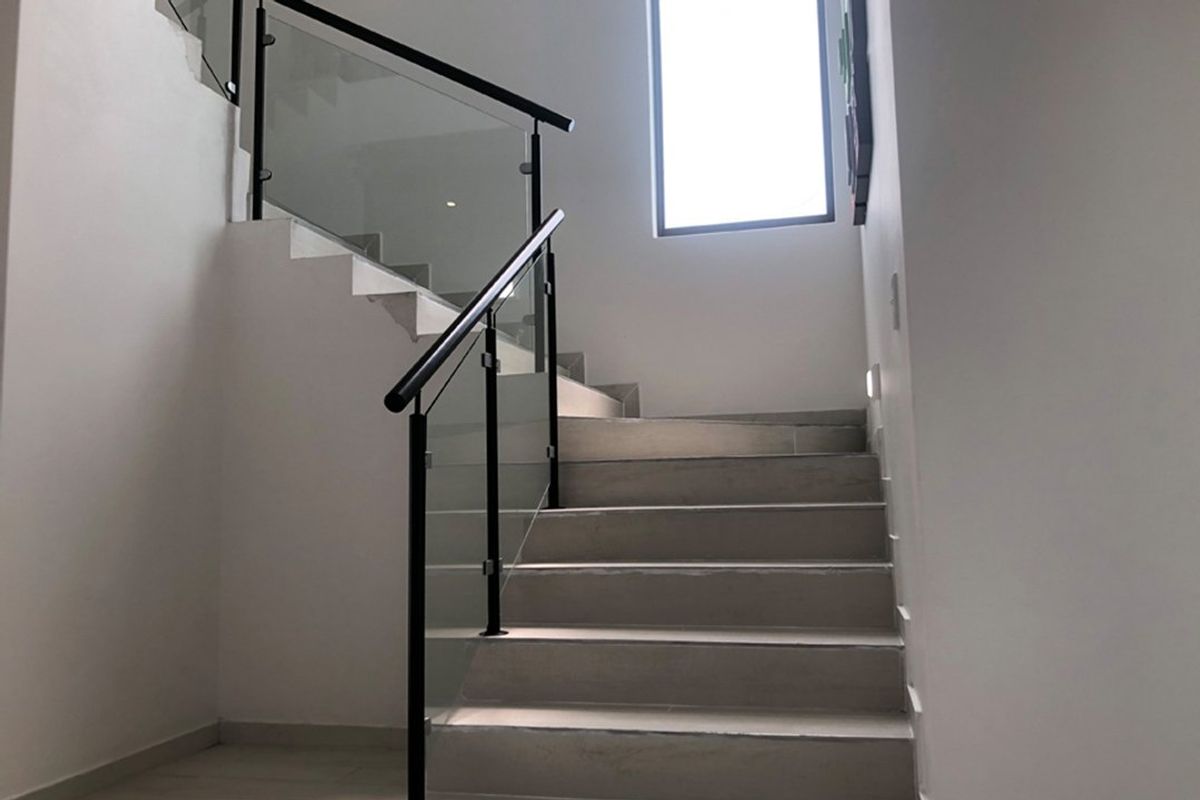 A modern staircase featuring tiled steps, a sleek glass balustrade, and a black metal handrail, illuminated by natural light from a large window.
