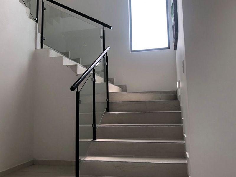 A modern staircase featuring tiled steps, a sleek glass balustrade, and a black metal handrail, illuminated by natural light from a large window.