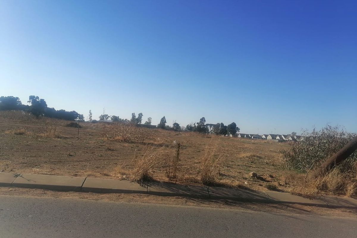 A view of a vacant residential plot from the adjacent asphalt road, featuring a concrete curb and surrounding landscape.