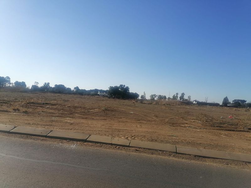 A vacant residential plot of land featuring level terrain, accessible from an adjacent asphalt road with established concrete kerbing.