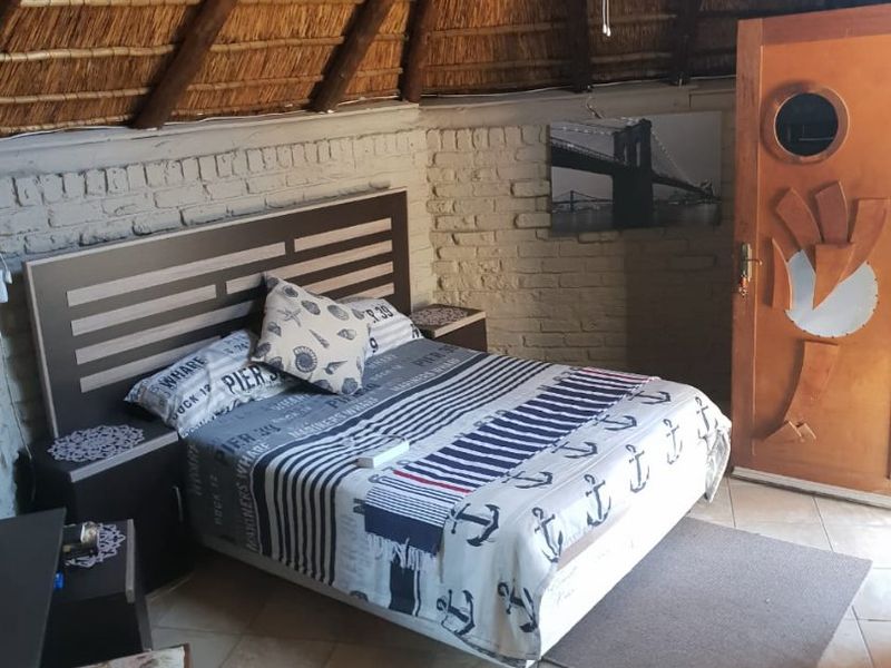 A rustic bedroom featuring a high thatched roof with exposed timber beams, tiled flooring, and a unique wooden door with a circular window.