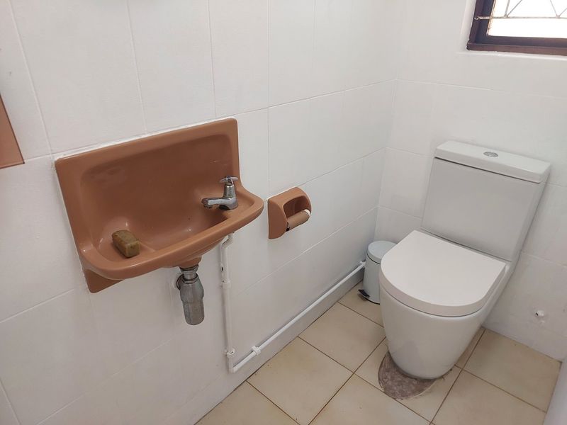 A compact bathroom featuring a wall-mounted basin, a standard toilet, and tiled finishes. The space is functional but would benefit from modern updates.