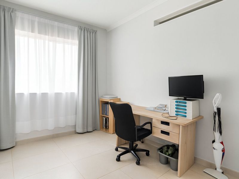 A bright and functional home office space featuring neutral tiled flooring, large window with curtains, and a wall-mounted air conditioning unit.
