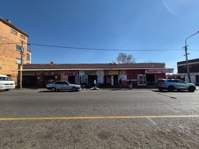 Street-level view of a commercial strip featuring multiple storefronts with metal security gates and a continuous corrugated metal awning.