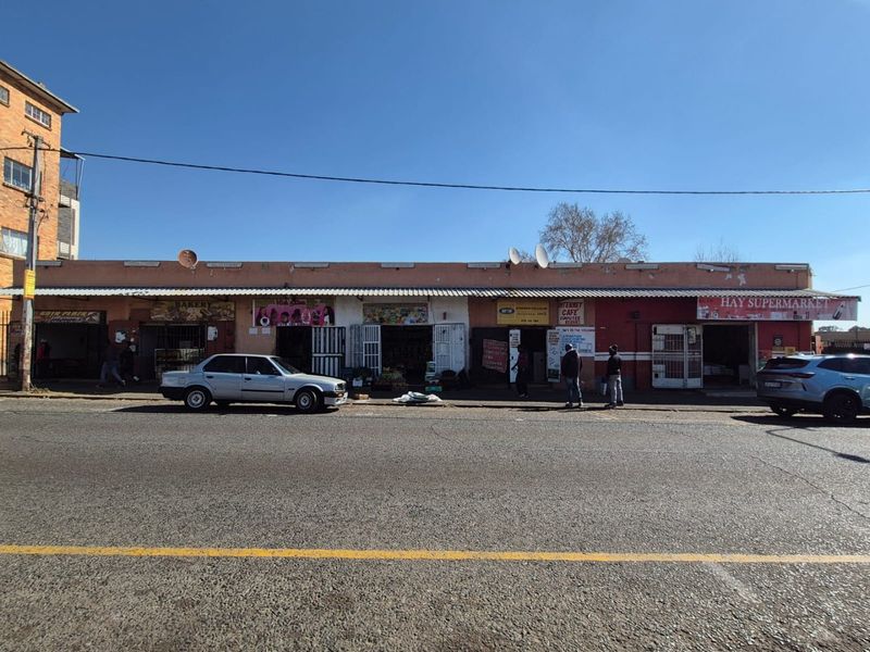 Street-level view of a commercial strip mall featuring multiple storefronts with security gates and a continuous corrugated metal awning.