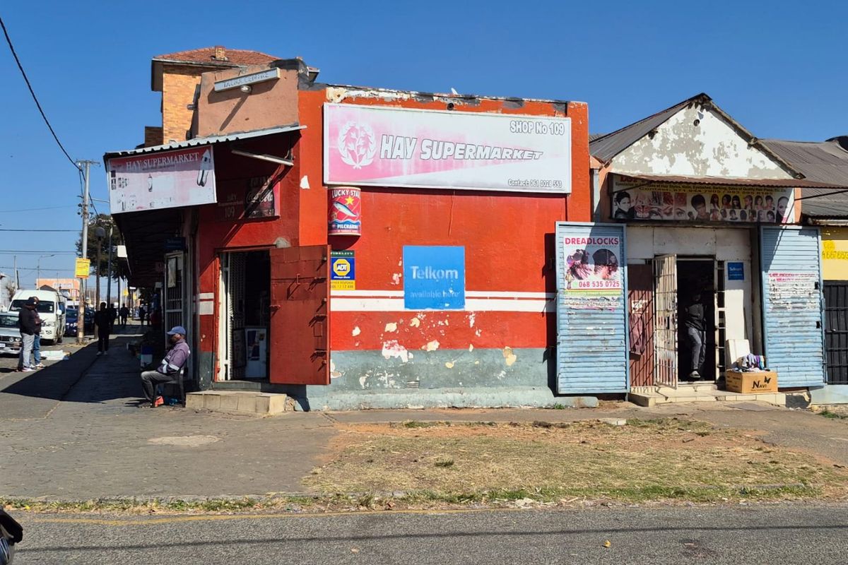 Street-level view of a commercial building featuring multiple shop fronts with security gates and signage in a high-traffic urban area.