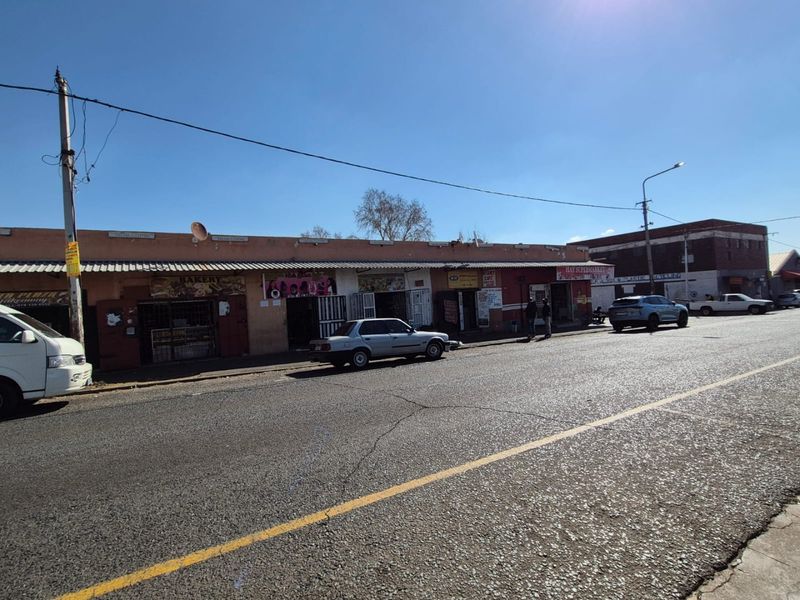 Street-level view of a commercial strip center featuring multiple storefronts with security gates and a corrugated metal roof.