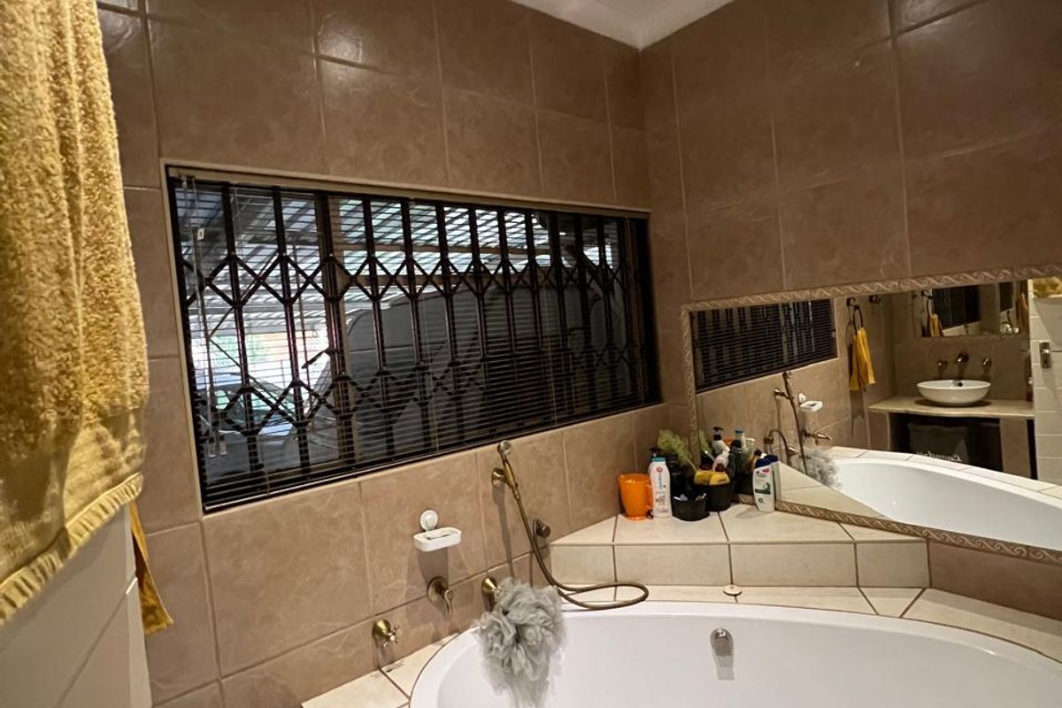 A bathroom featuring a built-in corner bathtub with tiled surround, a large wall mirror, and a window fitted with security bars.