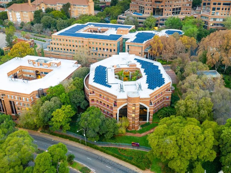 Aerial view of a commercial office complex featuring brick buildings with rooftop solar panels, surrounded by mature trees and landscaped grounds.