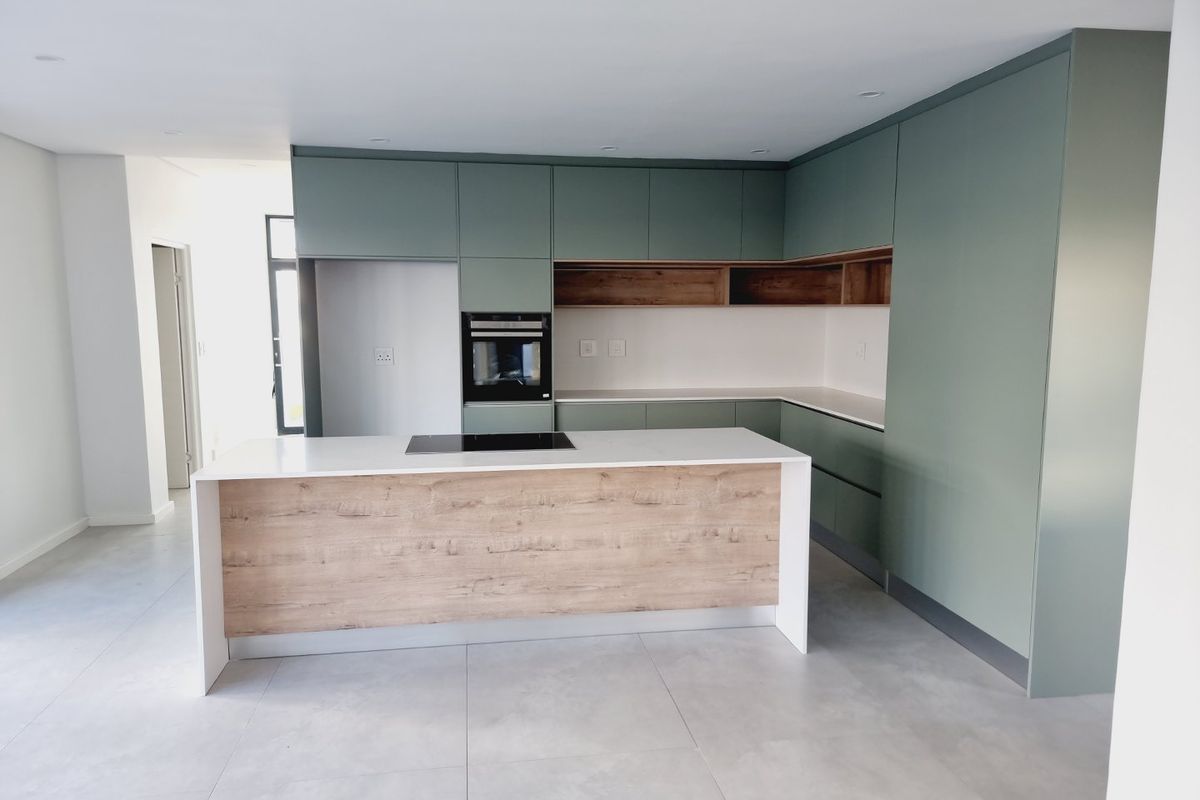 Modern kitchen featuring sage green cabinetry, wood-look accents, a central island with an integrated cooktop, and sleek white countertops.