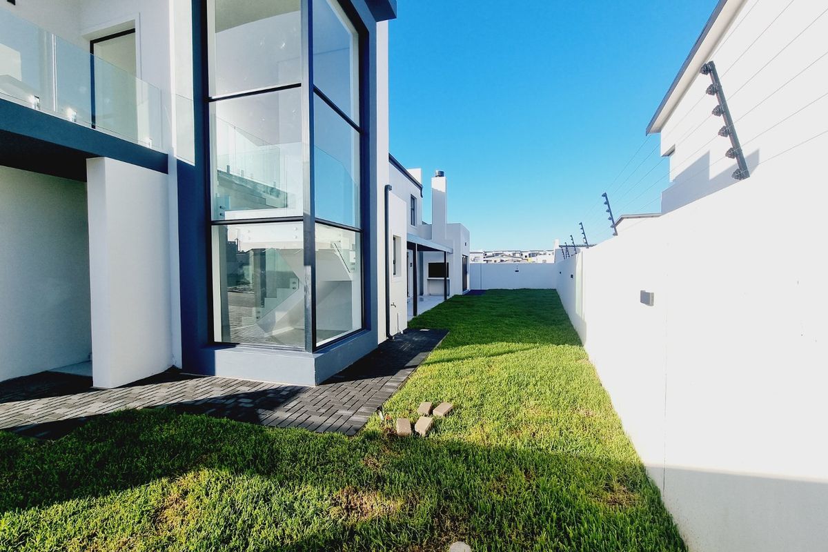 A modern rear garden area featuring a manicured lawn, paved walkway, and a striking double-volume glass facade with contemporary architectural design.