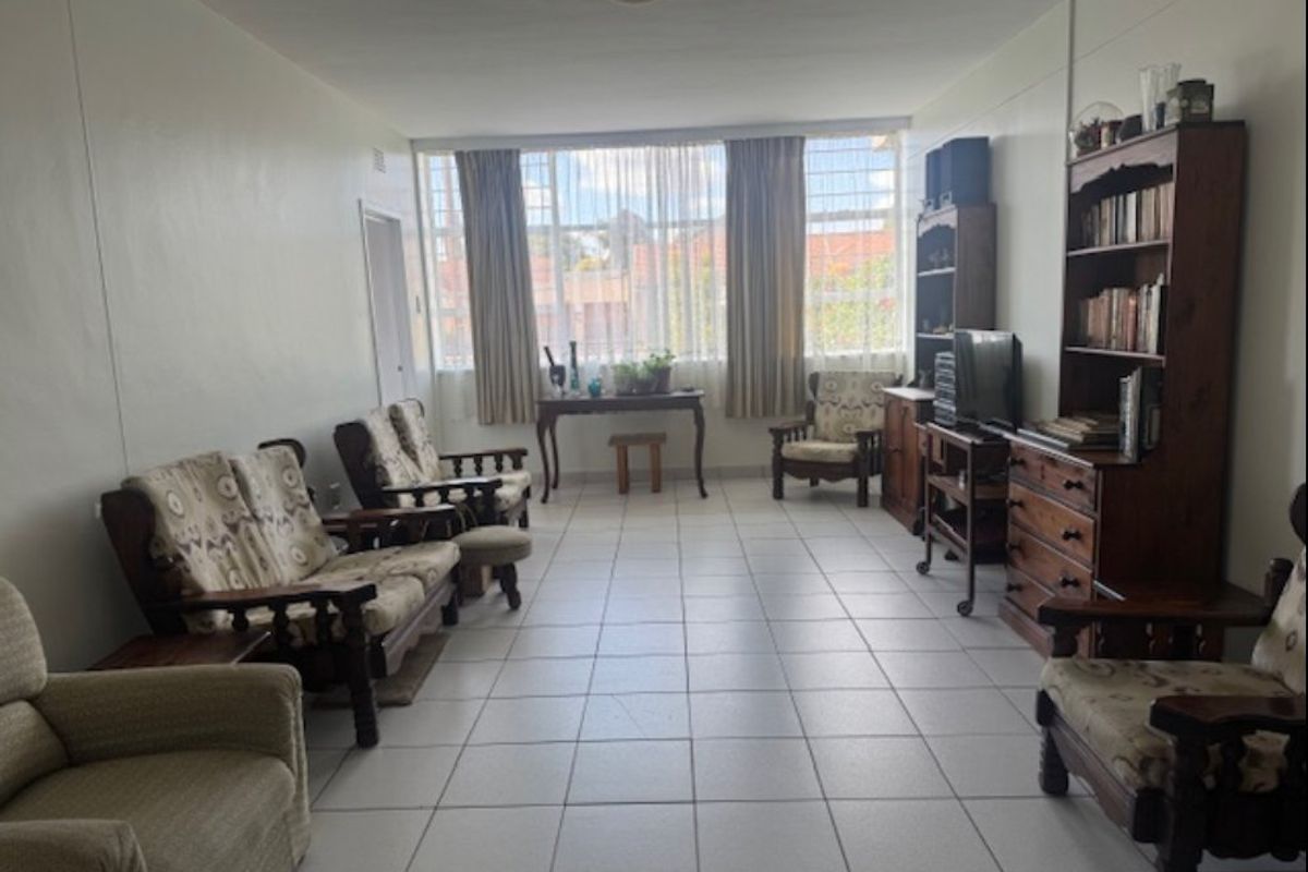 A spacious living room featuring neutral tiled flooring and a large window with security bars, offering a bright and functional living area.