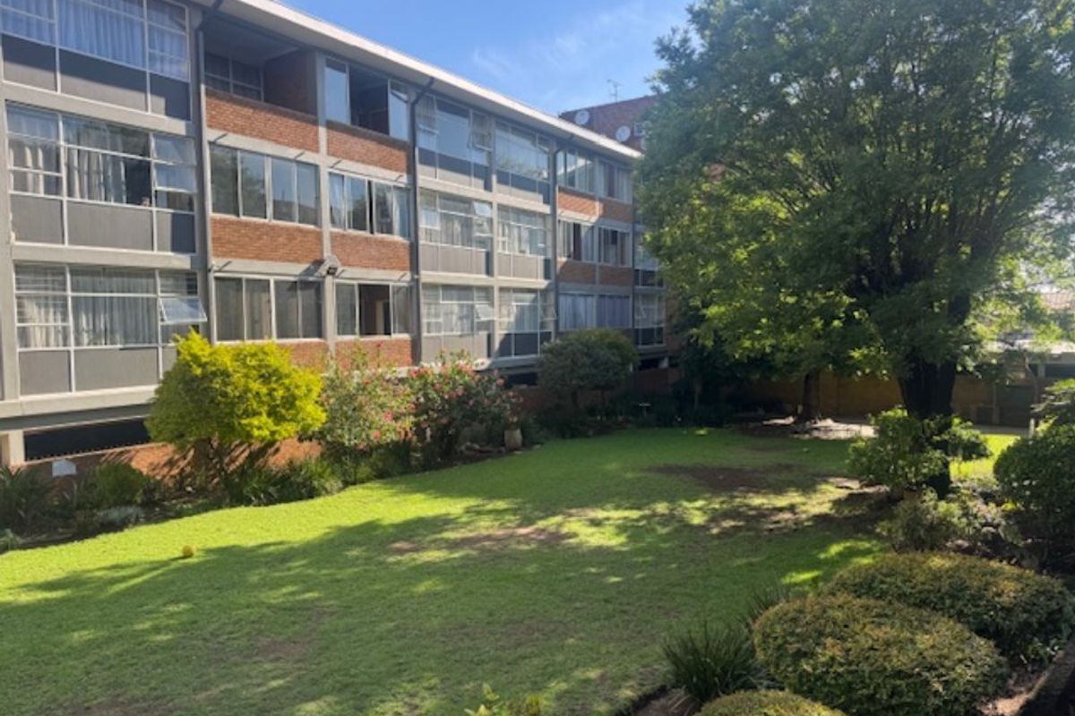 A view of the rear apartment building facade overlooking a well-maintained communal garden area with mature trees and lawn.