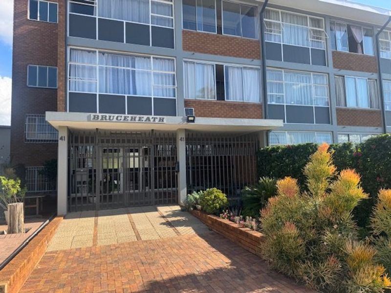 Front entrance view of the Bruceheath apartment complex featuring a brick facade, secure gated access, and a paved driveway with landscaped garden beds.