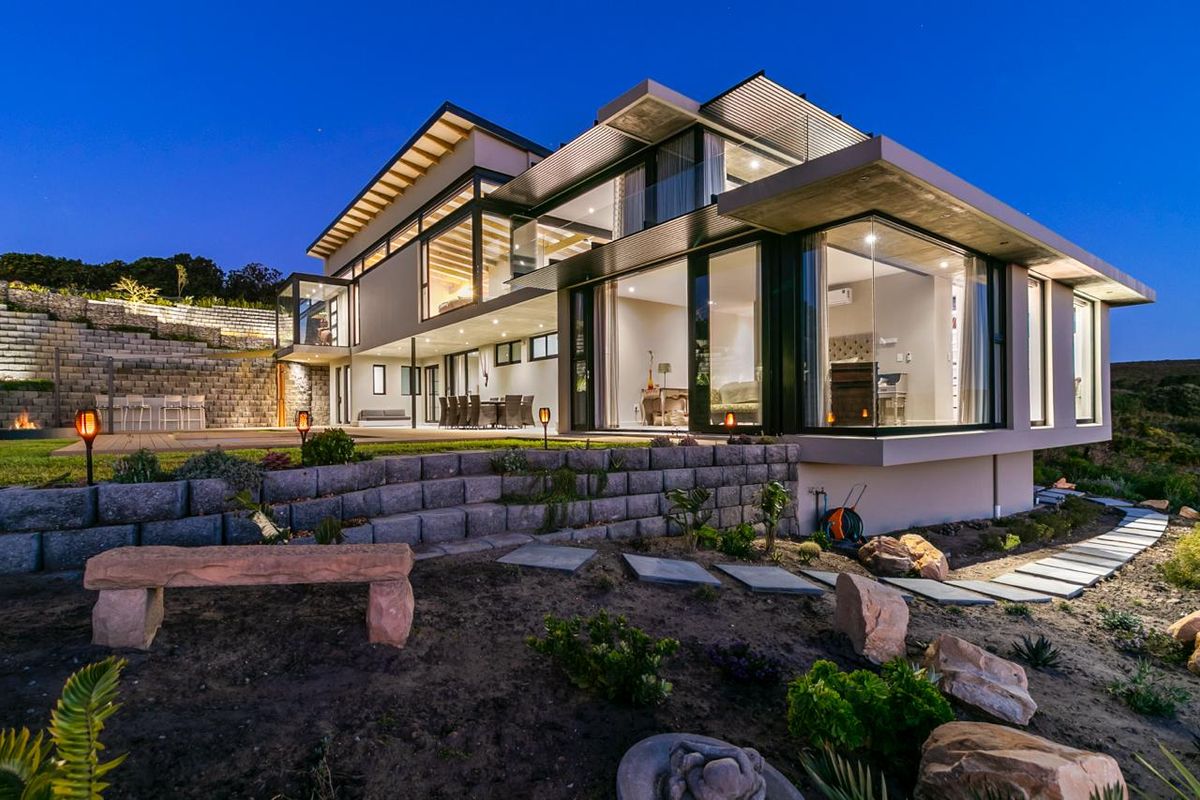 A stunning modern multi-level residence featuring expansive glass walls, cantilevered architecture, and tiered stone retaining walls, beautifully illuminated at dusk.