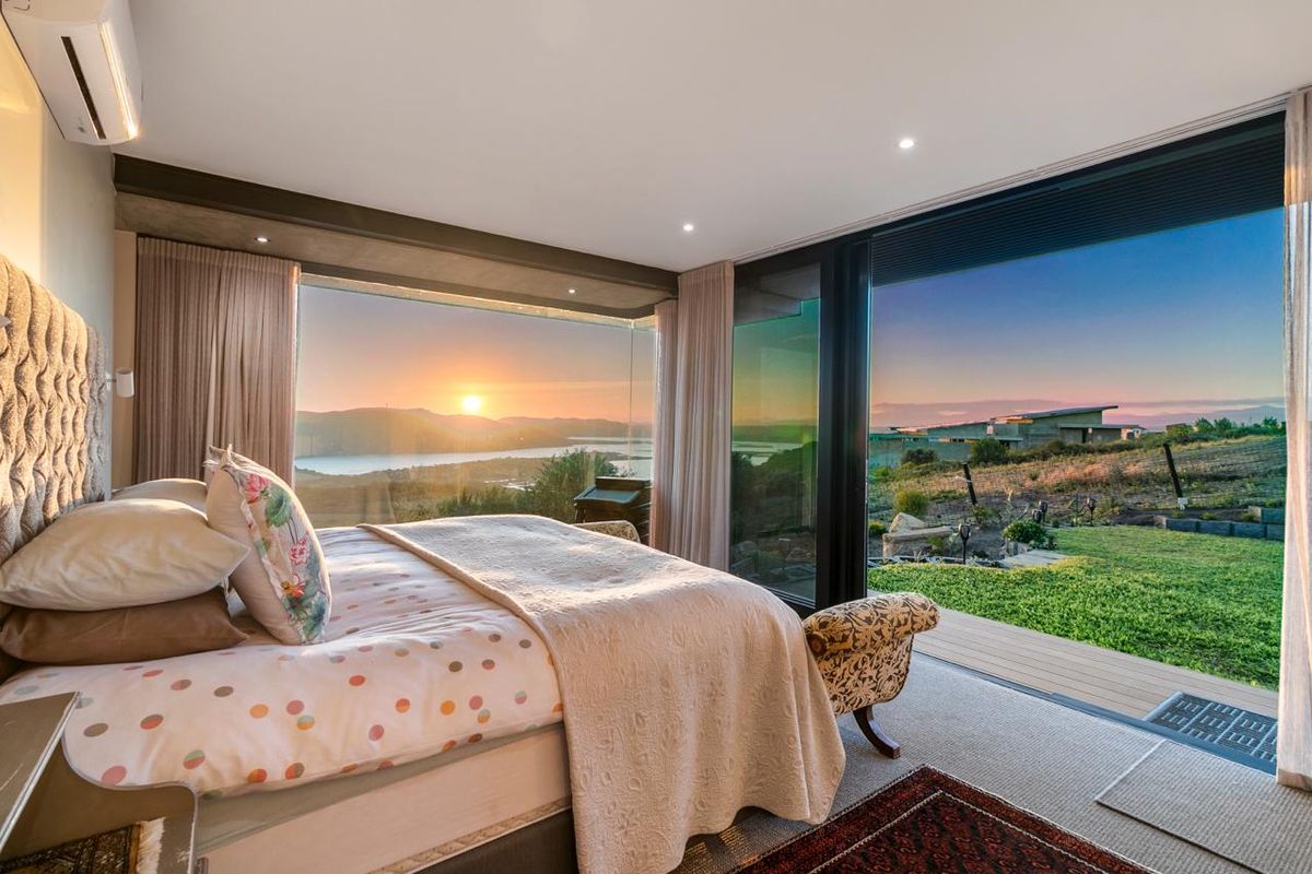 A modern bedroom featuring floor-to-ceiling glass sliding doors that open to a deck, offering expansive sunset views over the landscape.
