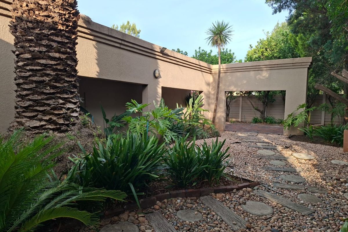 A rear garden area featuring a covered patio structure, stone pathway, and established landscaping with mature trees and shrubs.