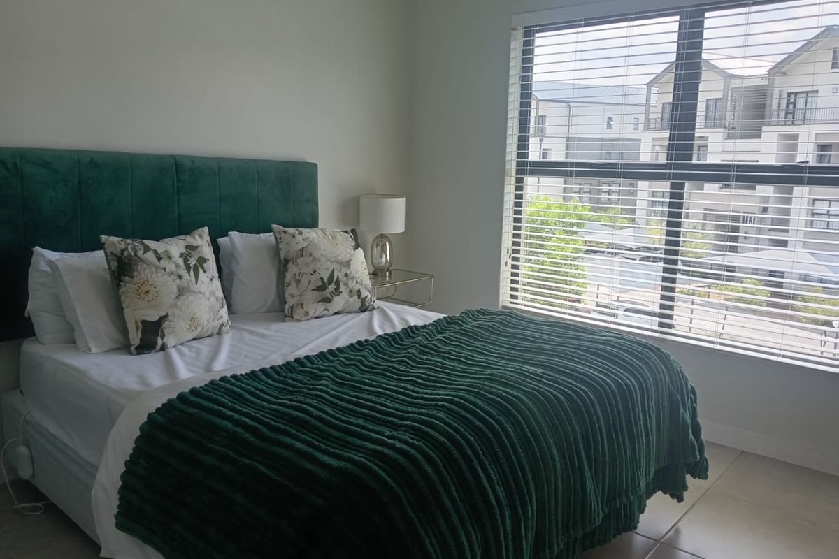 A well-lit bedroom featuring neutral tiled flooring and large windows fitted with venetian blinds, offering a clean and comfortable living space.