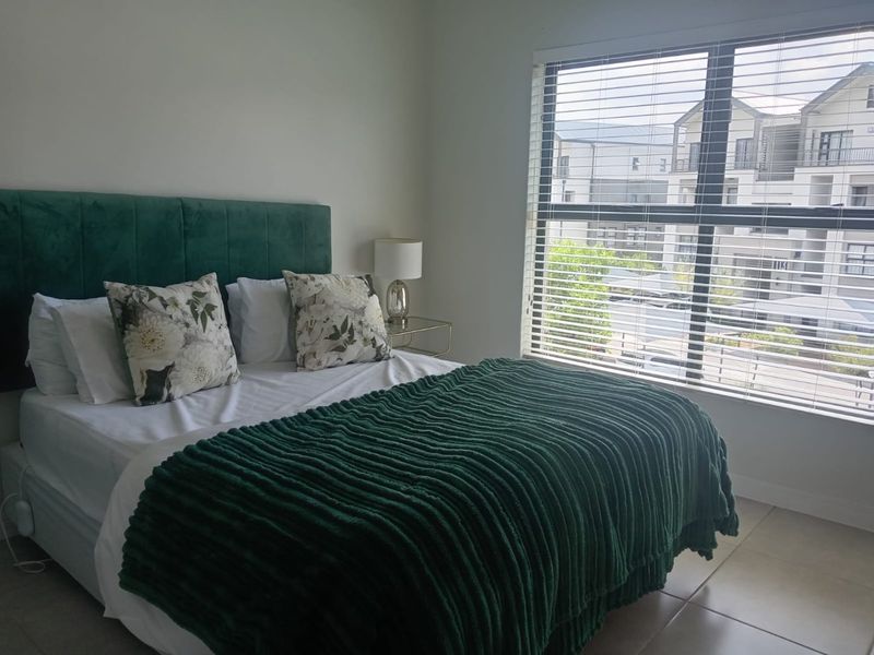 A well-lit bedroom featuring neutral tiled flooring and large windows fitted with venetian blinds, offering a clean and comfortable living space.