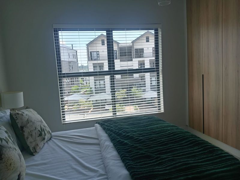 A well-maintained bedroom featuring modern built-in wooden cupboards and a large window with venetian blinds, offering a view of the surrounding apartment complex.
