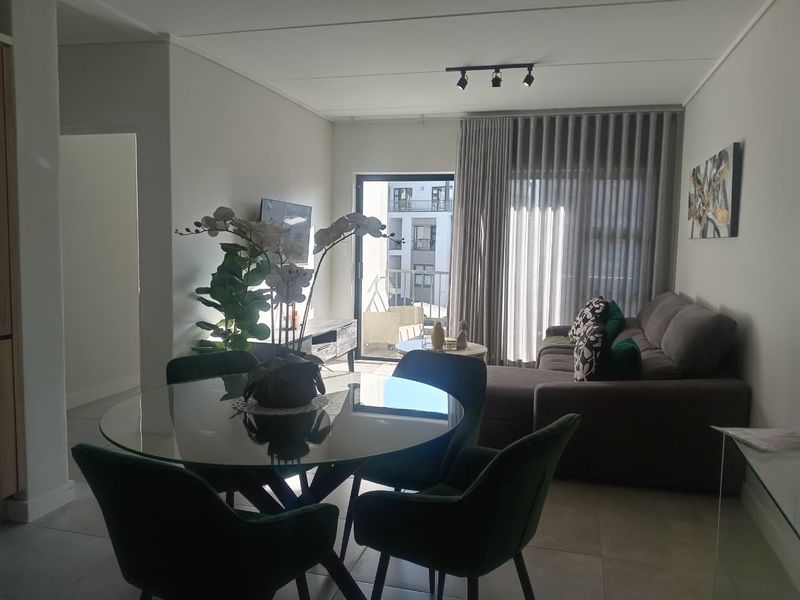 A bright open-plan living and dining area featuring tiled floors, modern track lighting, and a sliding glass door leading to a balcony.