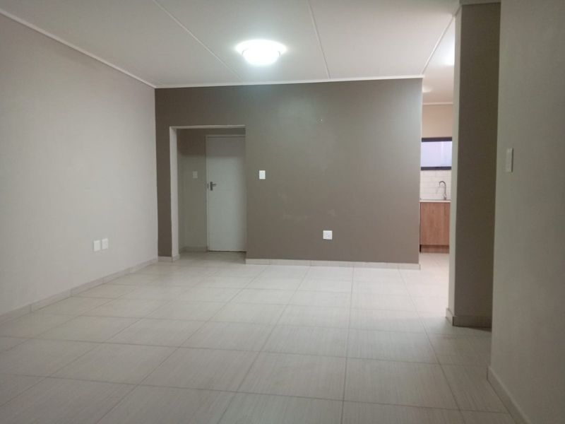 An open-plan living area featuring neutral tiled flooring, clean wall finishes, and a glimpse into the adjacent kitchen space.