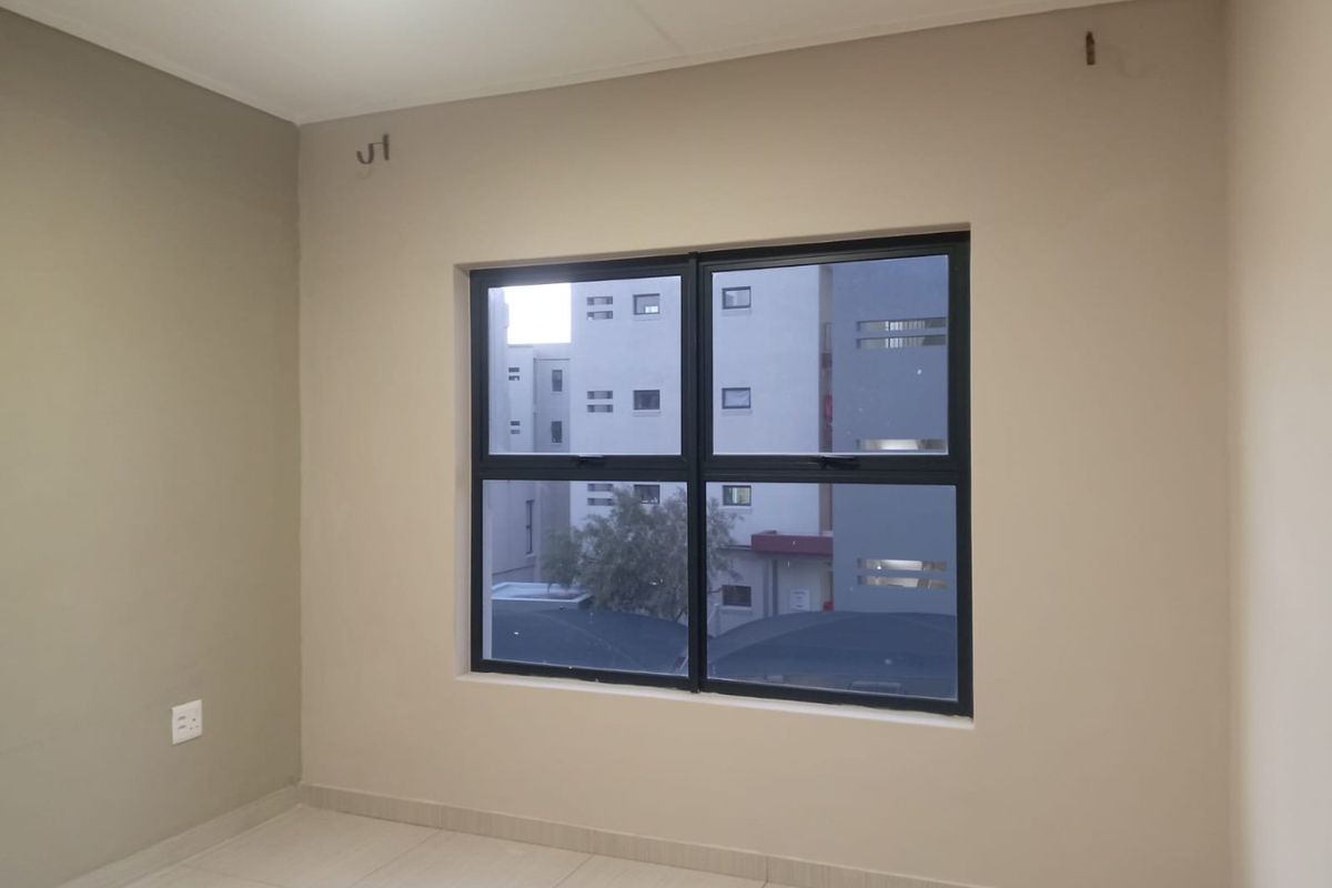 An empty bedroom featuring neutral painted walls, tiled flooring, and an aluminium-framed window with views of the surrounding apartment complex.
