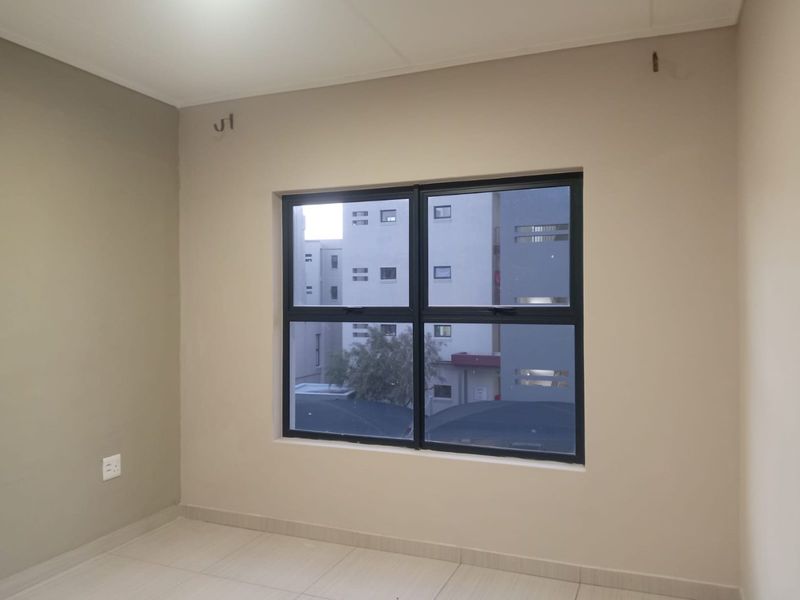 An empty bedroom featuring neutral painted walls, tiled flooring, and an aluminium-framed window with views of the surrounding apartment complex.