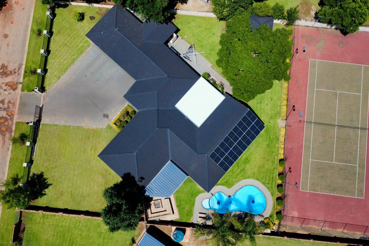 Aerial view of a residential property featuring a large tiled roof with solar panels, a swimming pool, and a private tennis court.