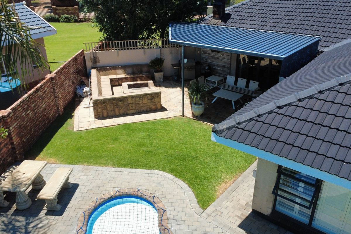 Aerial view of a rear garden featuring a paved patio, covered entertainment area with built-in braai, swimming pool, and well-maintained lawn.