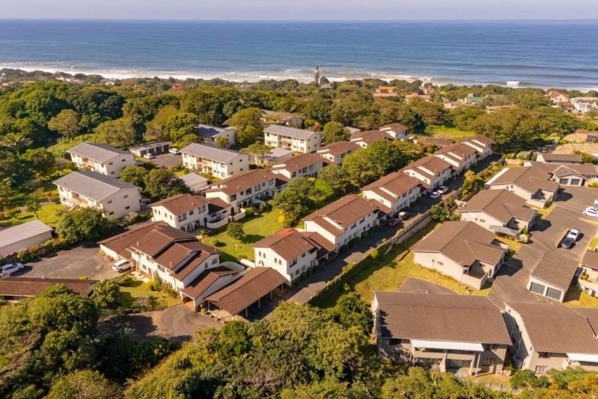 Aerial view of a coastal townhouse complex featuring multiple residential units, tiled roofs, paved driveways, and established communal gardens near the ocean.