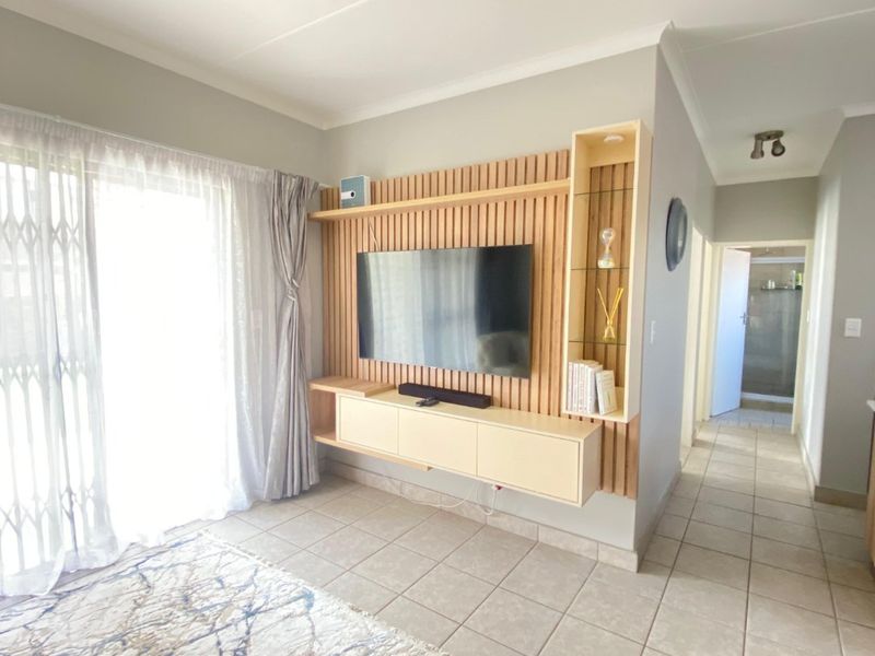 A bright living room featuring a modern built-in media unit with timber slat detailing, ceramic tiled flooring, and direct access to a balcony.
