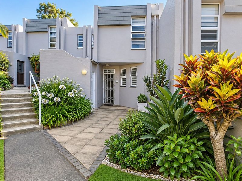 A well-maintained townhouse entrance featuring a paved walkway, concrete stairs with a handrail, and established garden landscaping leading to a secure front door.