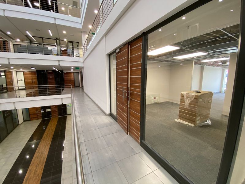 A modern commercial office hallway featuring glass partitions, wooden double doors, and a view overlooking a multi-level atrium with tiled flooring.