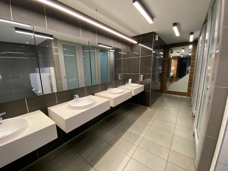 Modern commercial bathroom featuring multiple wall-mounted vanity basins, large mirrors, linear lighting, and tiled finishes in a clean, contemporary design.