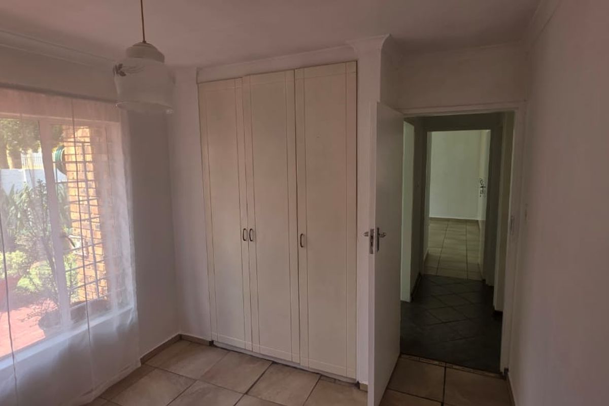 A bright, empty bedroom featuring built-in cupboards, tiled flooring, and a view through the window to the exterior area.