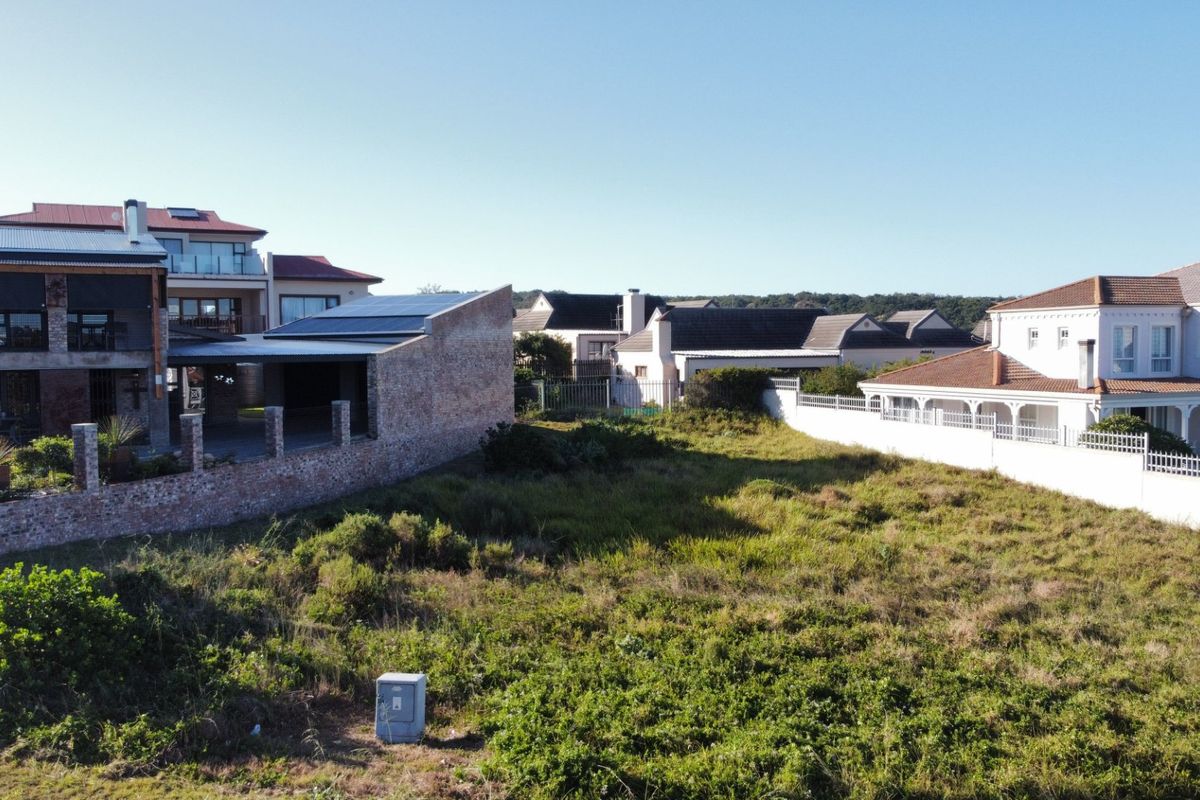 An elevated view of a vacant residential plot surrounded by established neighboring homes, featuring natural vegetation and boundary walls.