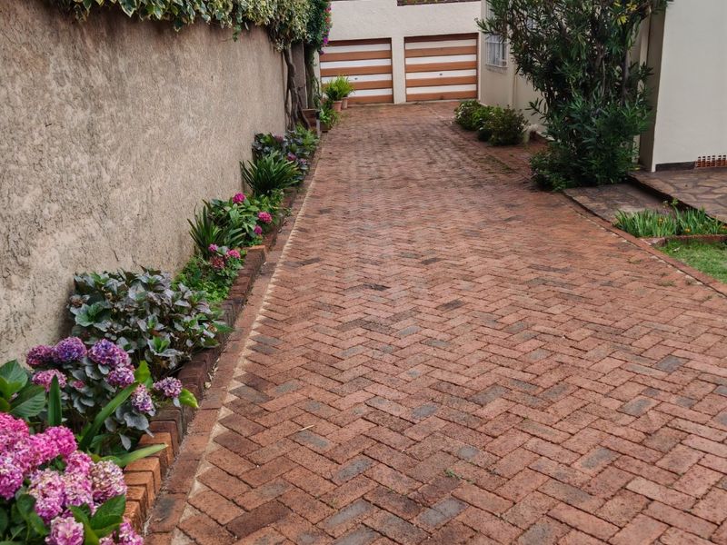 A well-maintained brick paved driveway leading to a double garage, flanked by a boundary wall and established garden beds.