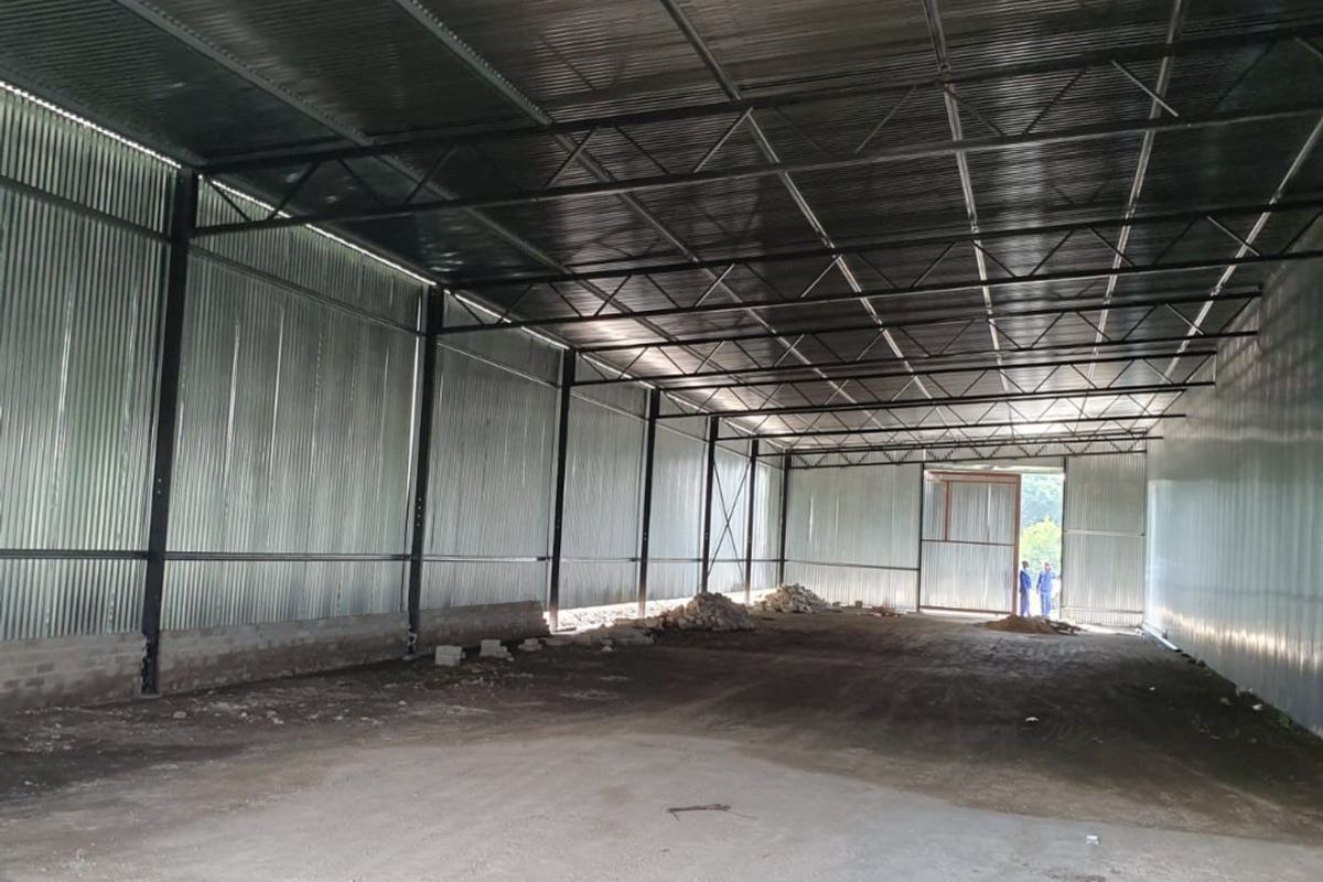 A large, open-plan industrial warehouse space featuring corrugated metal walls, exposed steel roof trusses, and a concrete floor with a large sliding entrance door.