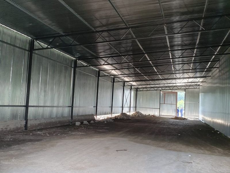 A large, open-plan industrial warehouse space featuring corrugated metal walls, exposed steel roof trusses, and a concrete floor with a large sliding entrance door.