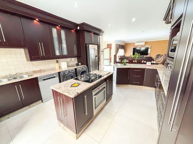 Spacious open-plan kitchen featuring dark wood cabinetry, granite countertops, a central island with a gas hob, and integrated appliances, flowing into the living area.