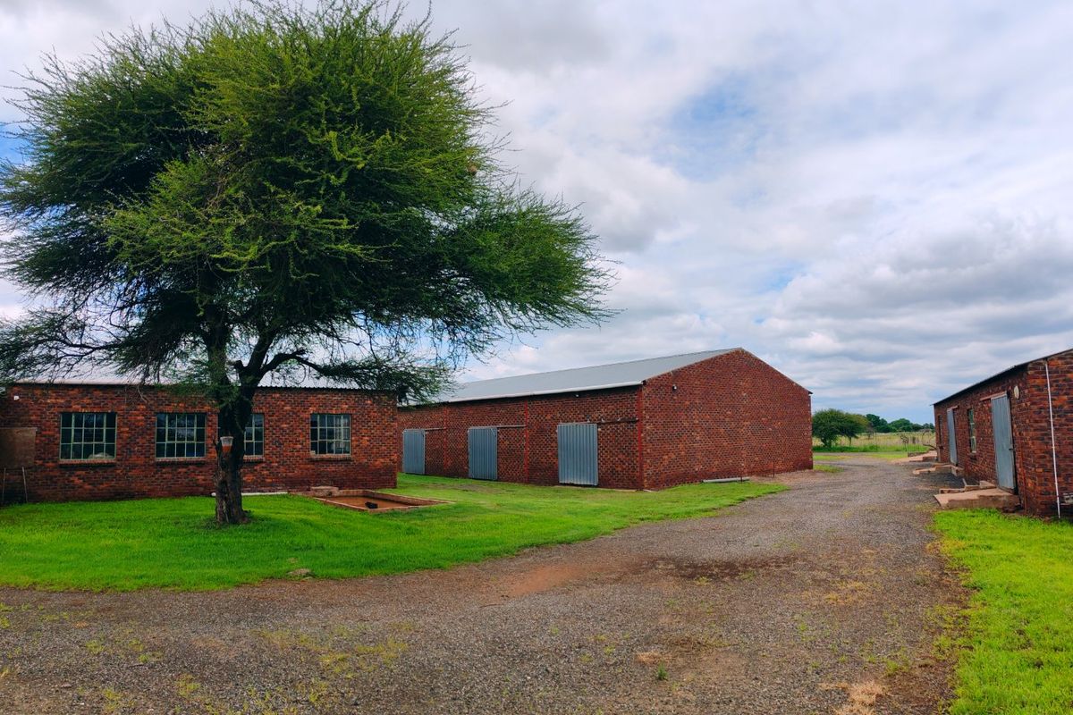 A view of multiple brick farm outbuildings with metal roofing, set around a gravel driveway and green lawn area.