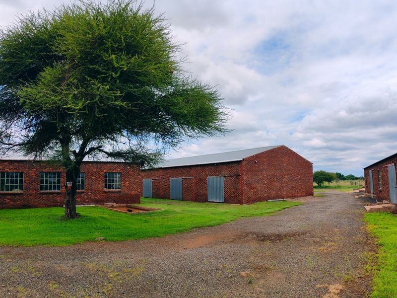 A view of multiple brick farm outbuildings with metal roofing, set around a gravel driveway and green lawn area.