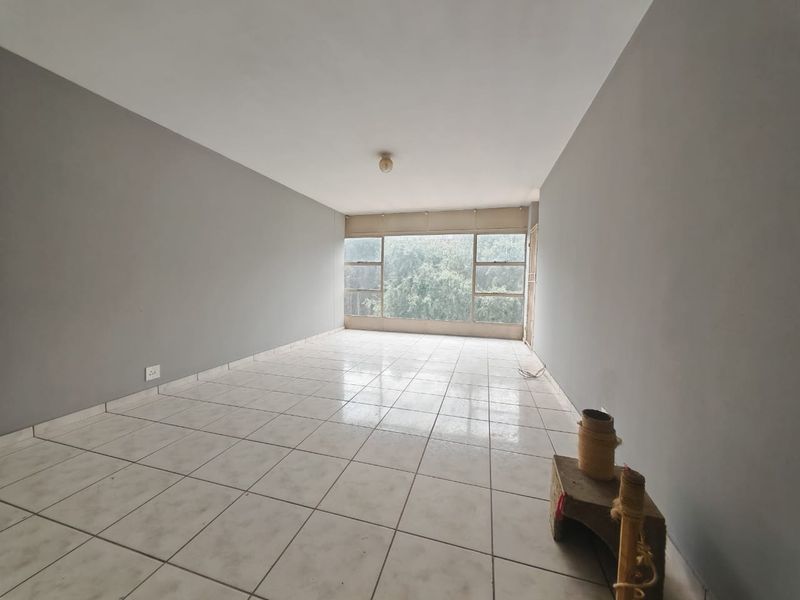 An empty, neutrally painted living area featuring large windows that provide natural light and tiled flooring throughout.