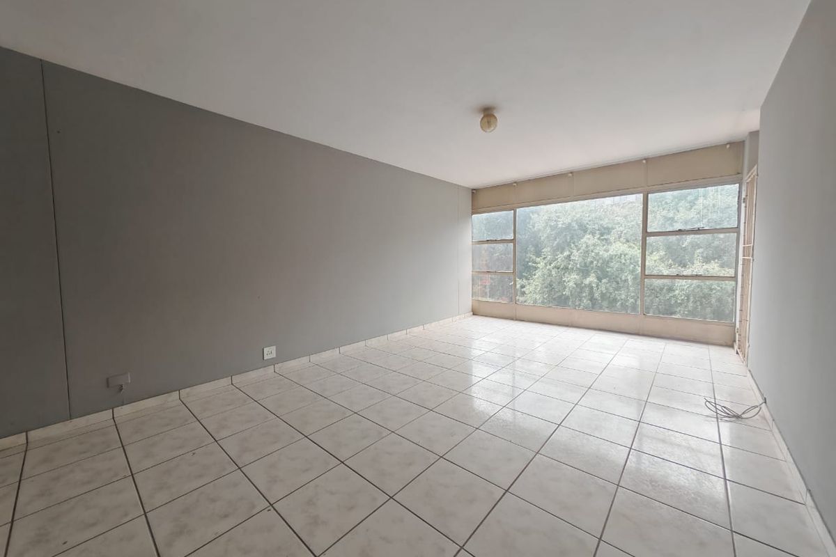An empty, sunlit living room featuring neutral grey walls, large windows with a garden view, and tiled flooring throughout.